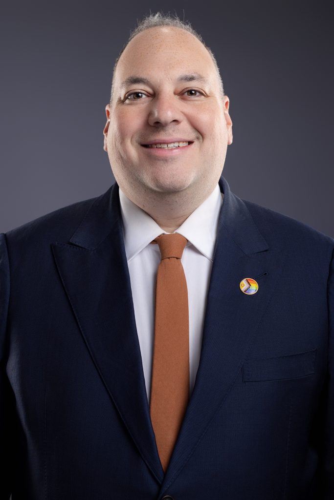 Image description: professional headshot of Adrian, a white man wearing a navy blue suit, white shirt, and orange tie. He is wearing an intersex-inclusive pride flag pin and standing in front of a grey background.